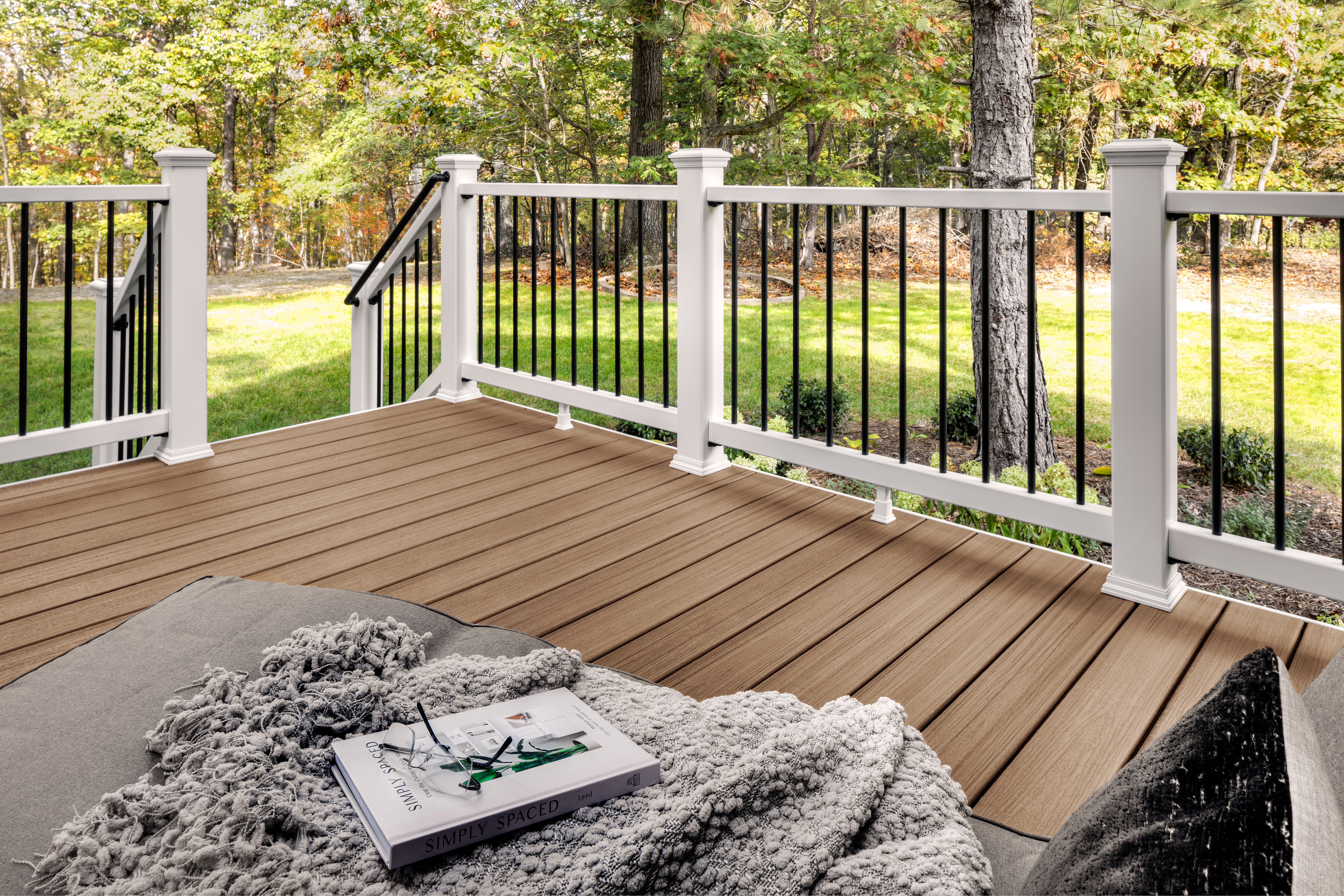 A wooden deck featuring a white and black railing surrounded by lush greenery. The setting includes a cozy blanket and a book placed on a soft cushion, suggesting relaxation. The scene is bright and inviting, with natural light filtering through the trees.